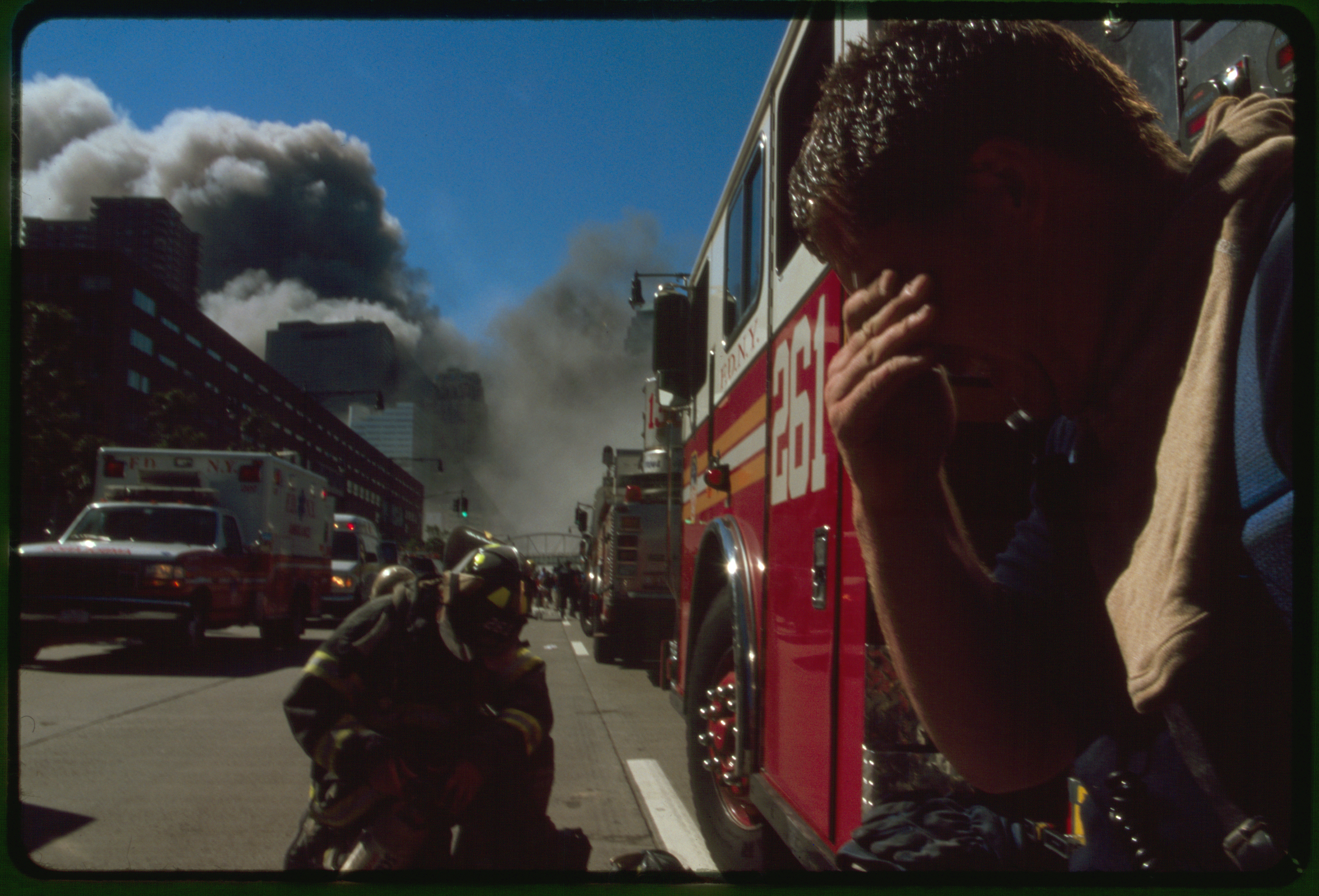 Fire fighter covering his eyes in foreground with New York City fire fighter, fire trucks, and rescue vehicles on street with burning buildings in background, following September 11th terrorist attack on World Trade Center, New York City