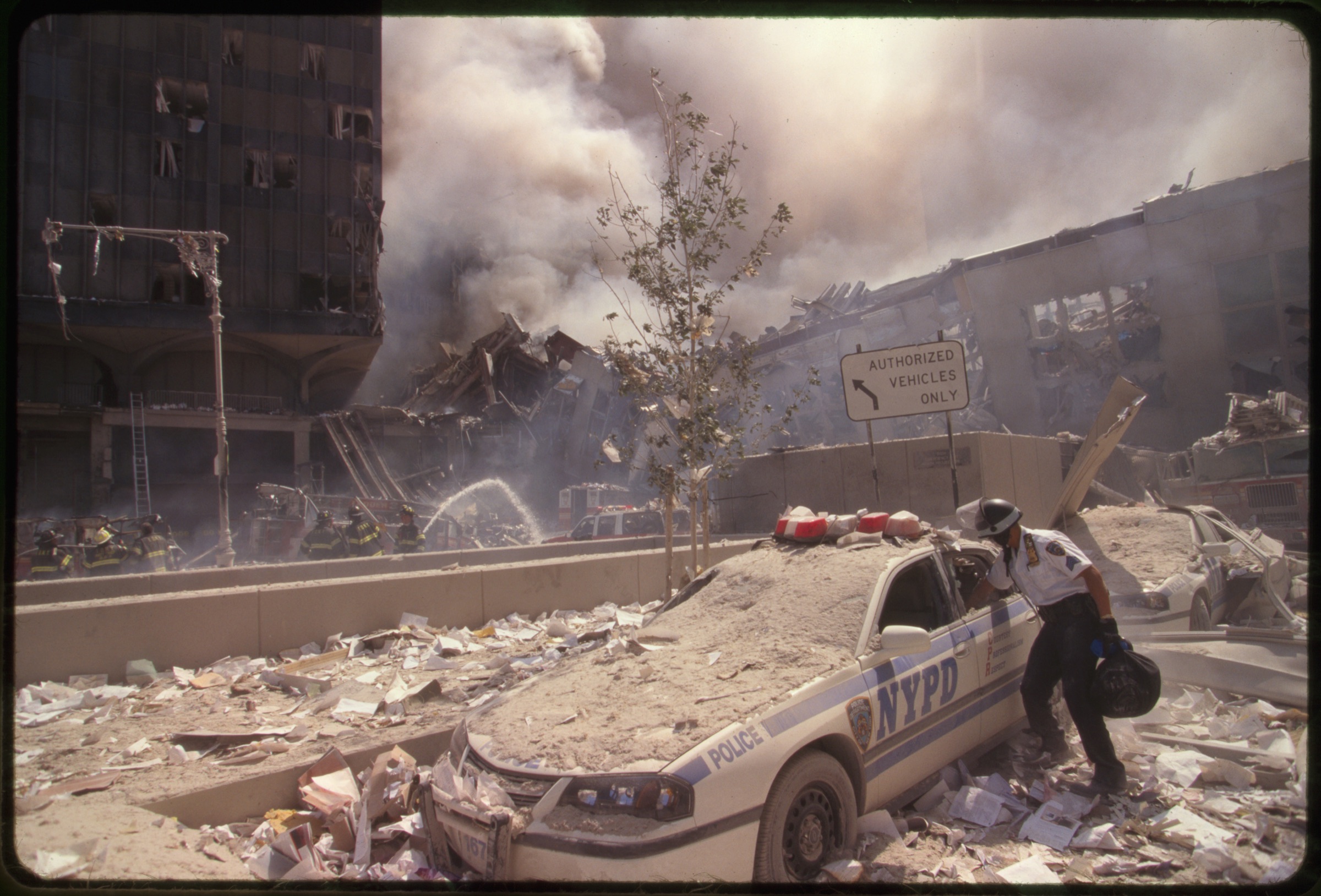 Rescue worker reaching into a New York Police car covered with debris while New York City fire fighters spray water on smoldering ruins in background, following September 11th terrorist attack on World Trade Center, New York City
