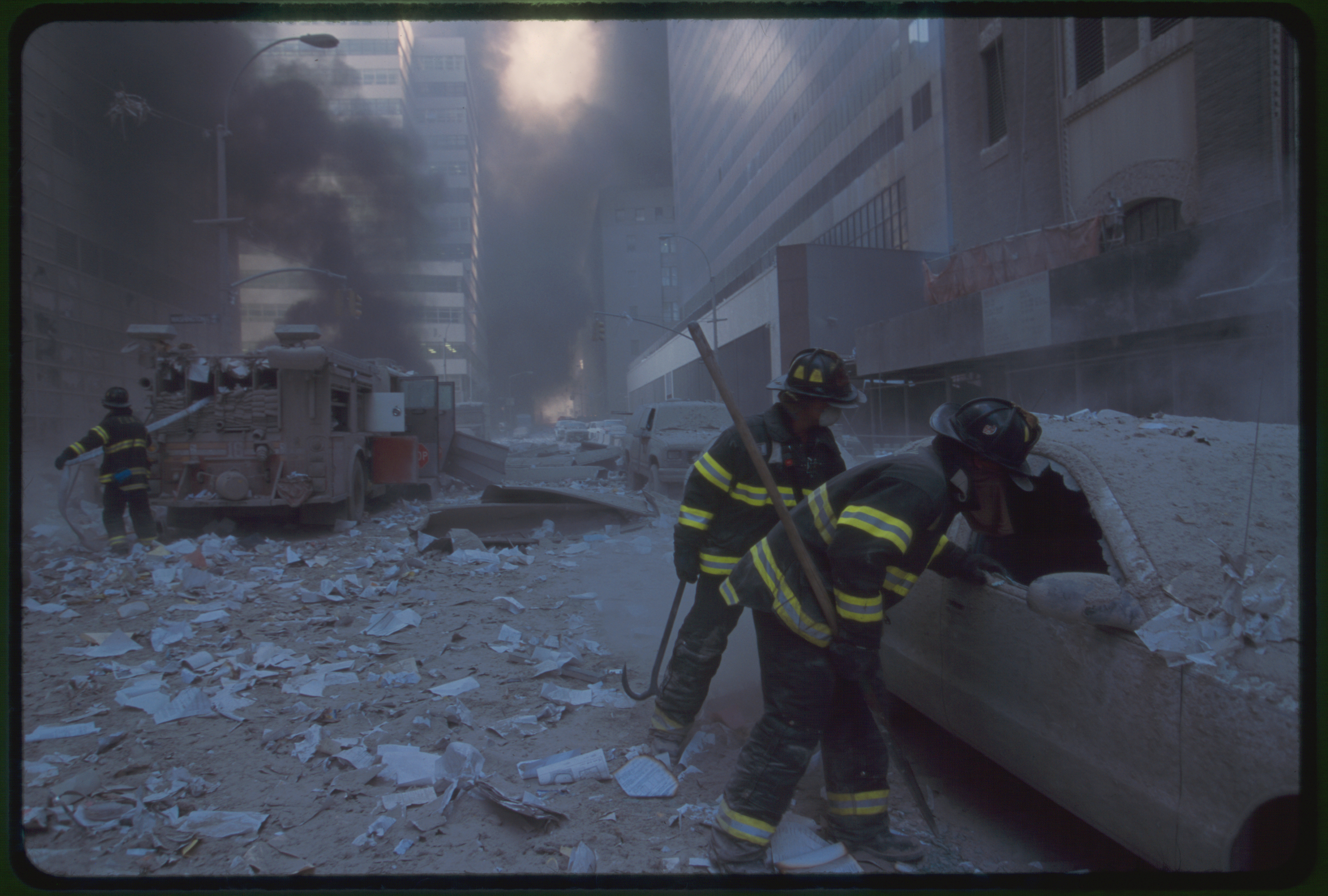 Two fire fighters looking into car while another New York City fire fighter pulls water hose from fire truck amid smoke and debris following September 11th terrorist attack on World Trade Center, New York City